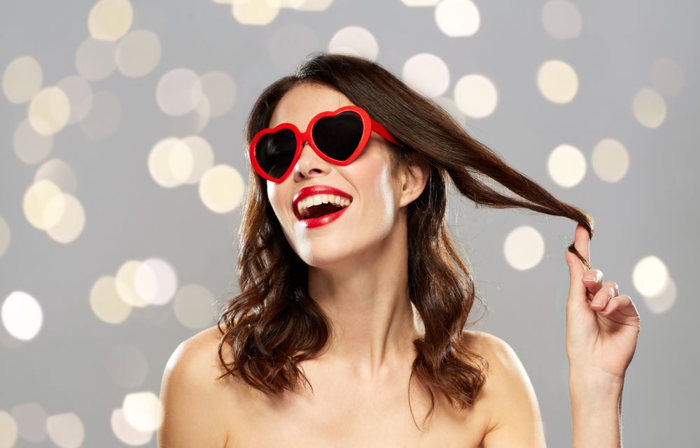 valentines day, holidays, beauty and people concept - happy smiling young woman with red lipstick and heart shaped sunglasses curling hair strand over gray background and lights. woman with red lipstick and heart shaped shades