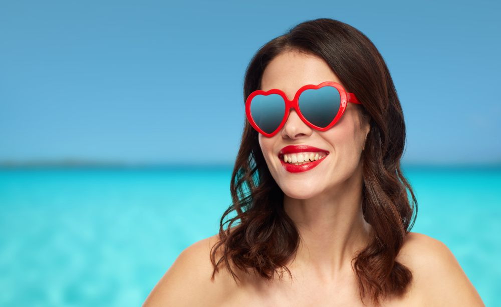 valentines day, beauty and people concept - happy smiling young woman with red lipstick and heart shaped sunglasses over ocean and blue sky background. woman with red lipstick and heart shaped shades