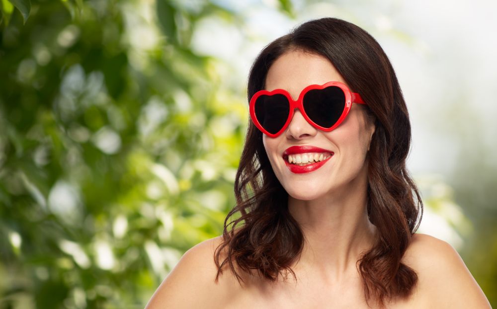 valentines day, beauty and people concept - happy smiling young woman with red lipstick and heart shaped sunglasses over green natural background. woman with red lipstick and heart shaped shades