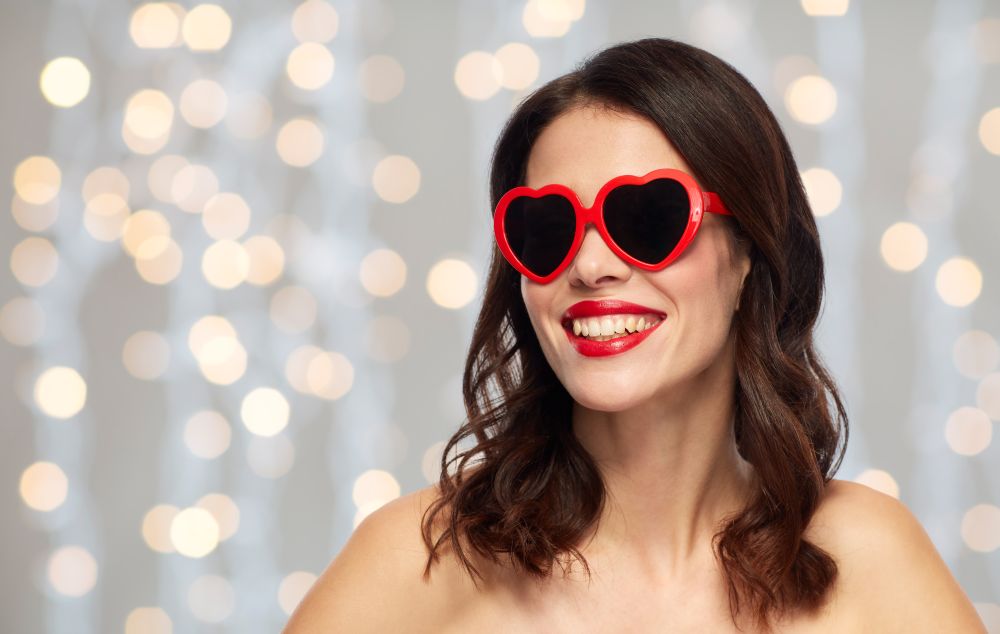 valentines day, beauty and people concept - happy smiling young woman with red lipstick and heart shaped sunglasses over holidays lights background. woman with red lipstick and heart shaped shades