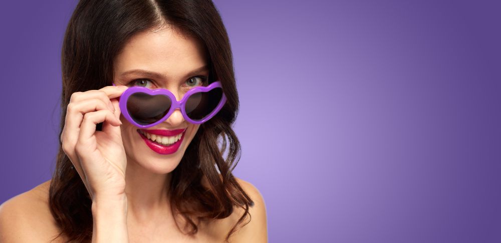 valentines day, beauty and people concept - close up of happy smiling young woman with lipstick and ultra violet heart shaped sunglasses. woman with ultra violet heart shaped shades