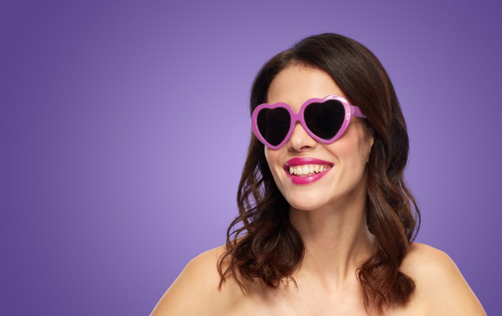 valentines day, beauty and people concept - close up of happy smiling young woman with berry lipstick and heart shaped sunglasses over ultra violet background. woman with heart shaped shades over ultra violet