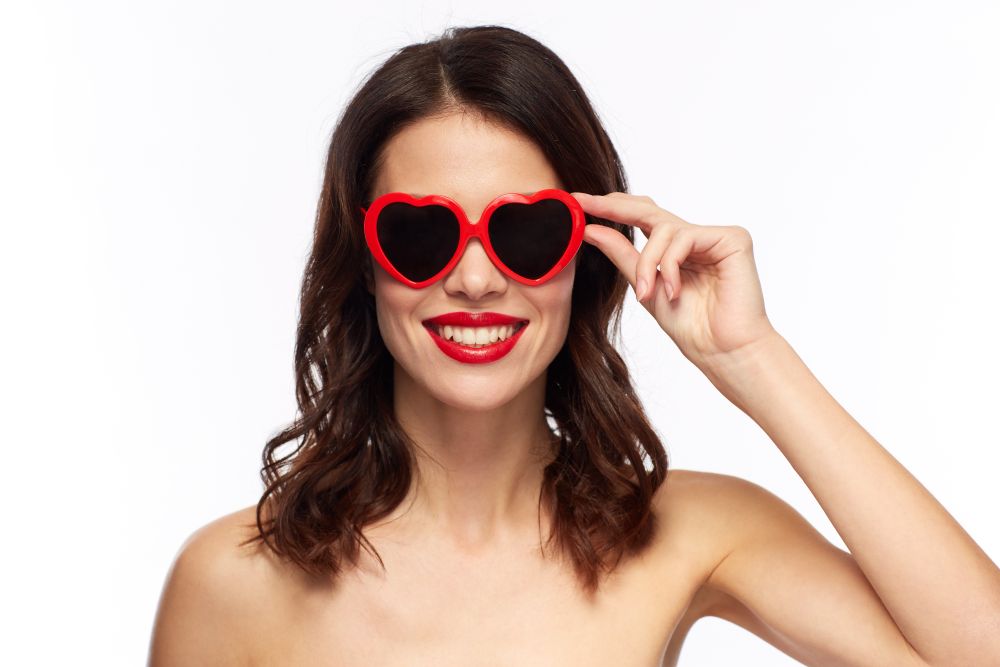 valentines day, beauty and people concept - happy smiling young woman with red lipstick and heart shaped sunglasses over white background. woman with red lipstick and heart shaped shades