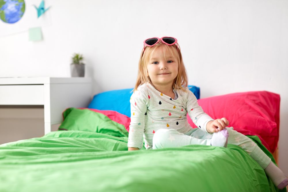 childhood and people concept - happy smiling little girl with sunglasses sitting on bed at home. smiling little girl with sunglasses on bed at home