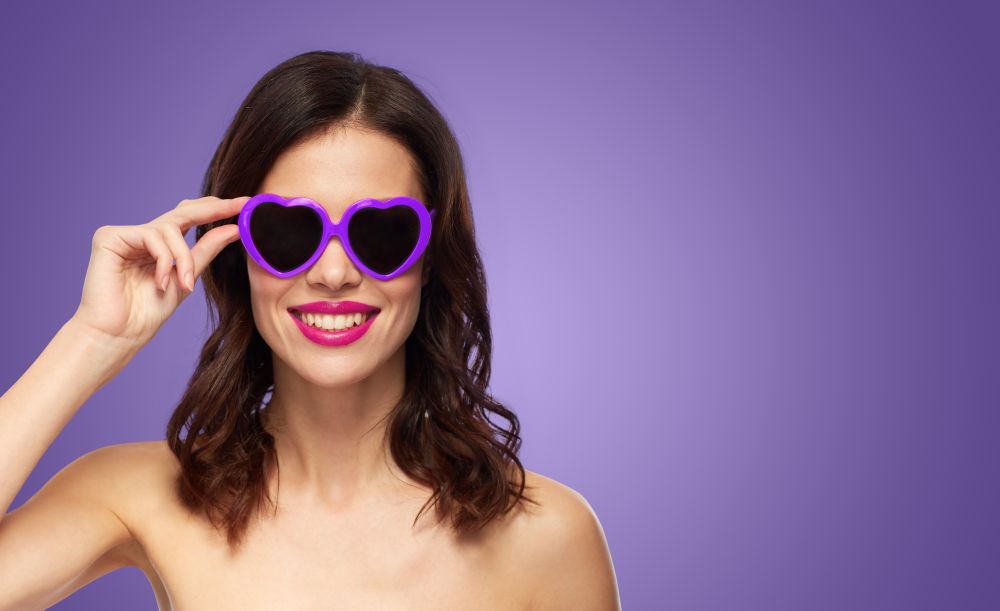 valentines day, beauty and people concept - happy smiling young woman with pink lipstick and heart shaped sunglasses over ultra violet background. woman with pink lipstick and heart shaped shades. woman with pink lipstick and heart shaped shades