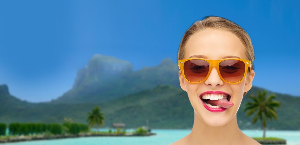 travel, tourism, summer holidays and vacation concept - smiling young woman in sunglasses with pink lipstick on lips over bora bora island beach background showing tongue. woman in shades showing tongue over summer beach. woman in shades showing tongue over summer beach