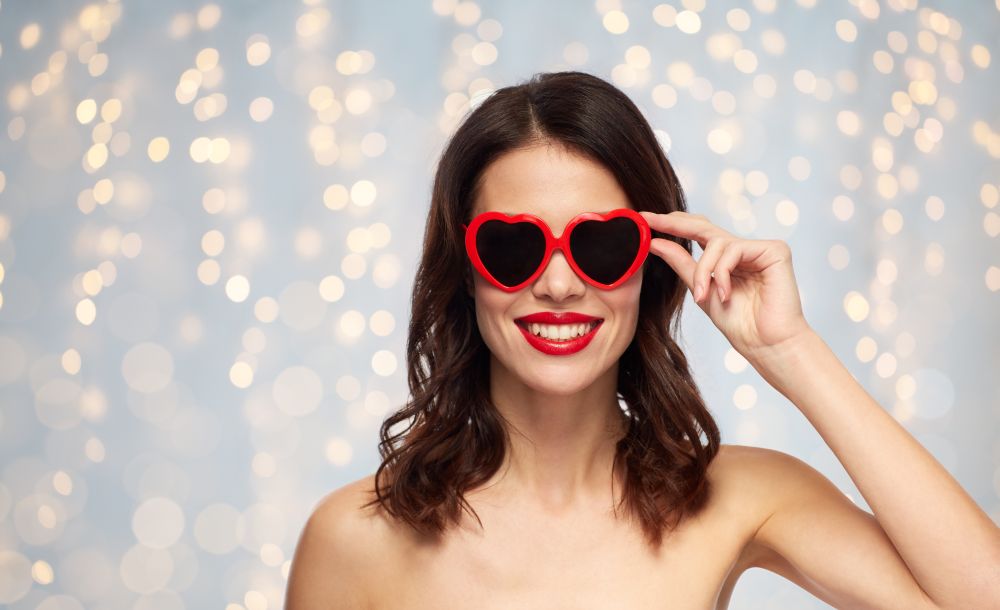 valentines day, beauty and people concept - happy smiling young woman with red lipstick and heart shaped sunglasses over holidays lights background. woman with red lipstick and heart shaped shades. woman with red lipstick and heart shaped shades