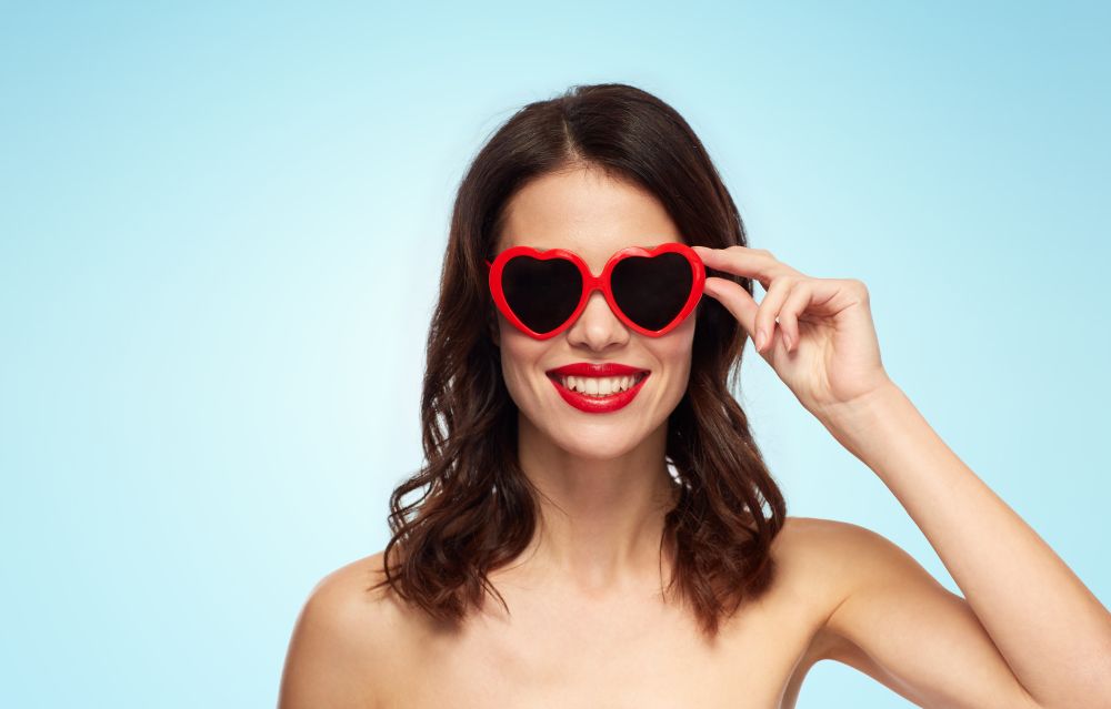 valentines day, beauty and people concept - happy smiling young woman with red lipstick and heart shaped sunglasses over blue background. woman with red lipstick and heart shaped shades. woman with red lipstick and heart shaped shades