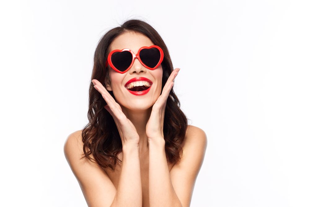valentines day, beauty and people concept - happy smiling young woman with red lipstick and heart shaped sunglasses over white background. woman with red lipstick and heart shaped shades. woman with red lipstick and heart shaped shades
