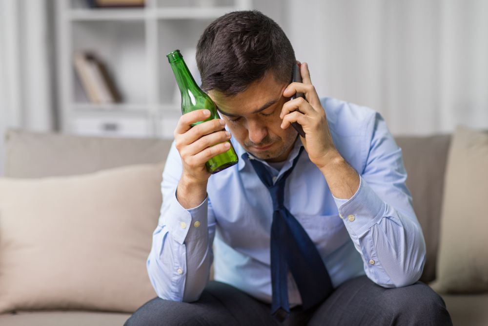 alcoholism, alcohol addiction and people concept - male alcoholic drinking beer and calling on smartphone at home. man drinking alcohol and calling on smartphone. man drinking alcohol and calling on smartphone