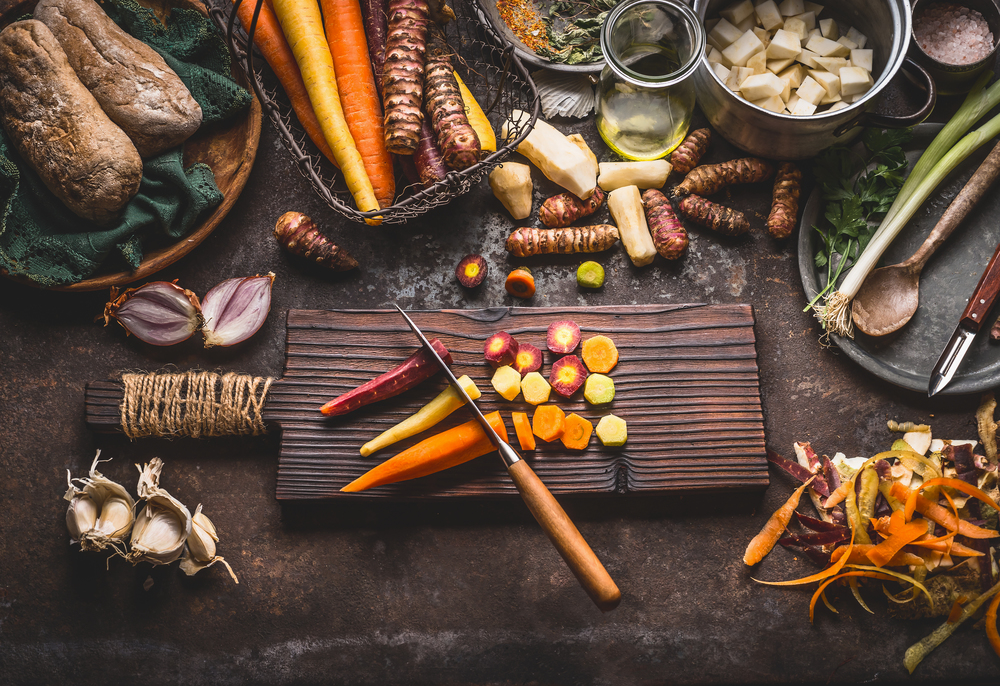 Colorful sliced carrots with knife on wooden cutting board on rustic kitchen table background with root vegetables ingredients for tasty vegetarian cooking, top view.  Healthy food and eating
