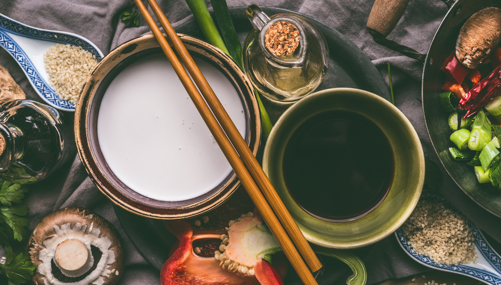 Bowls with coconut milk, soy sauce, sesame oil, chopsticks and vegetables on kitchen table background, top view.  Asian food cooking ingredients, Chinese or Thai cuisine concept