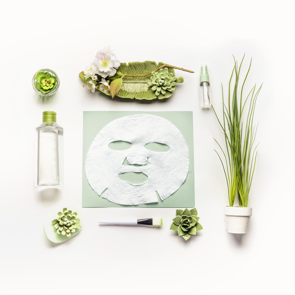 Modern facial skin care setting . Herbal cosmetic concept. Sheet mask with green cosmetic products, accessories plants and flowers on white desktop background, top view, copy space, flat lay.