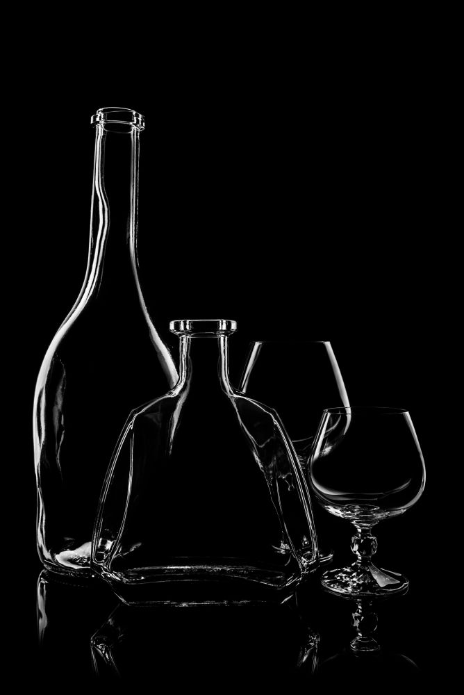 transparent bottle of brandy with glasses on black background with reflection