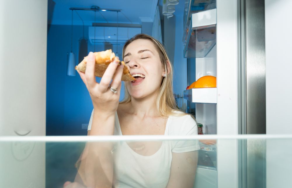 Portrait of beautiful woman eating pizza. View from inside of the fridge