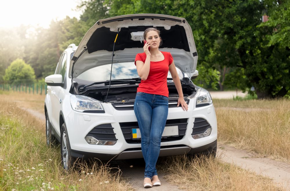 Young woman standing at broken car and talking by phone