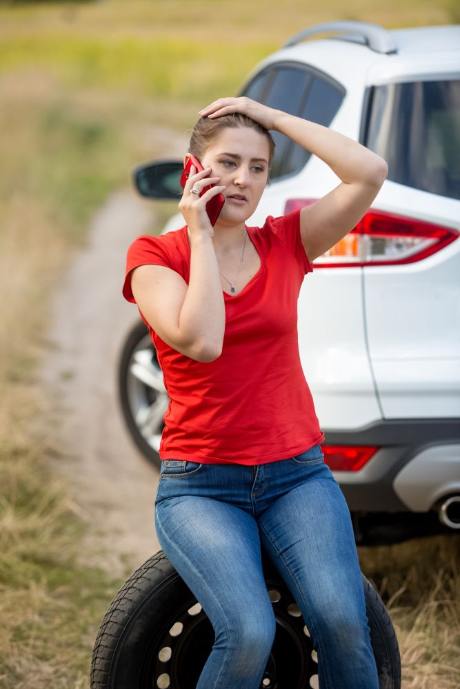 Portrait of young female driver sitting next broken car and talking by phone