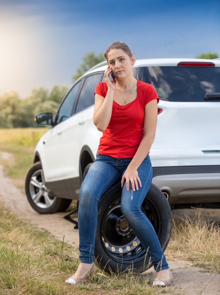 Young female driver sitting next broken car at field and talking by phone