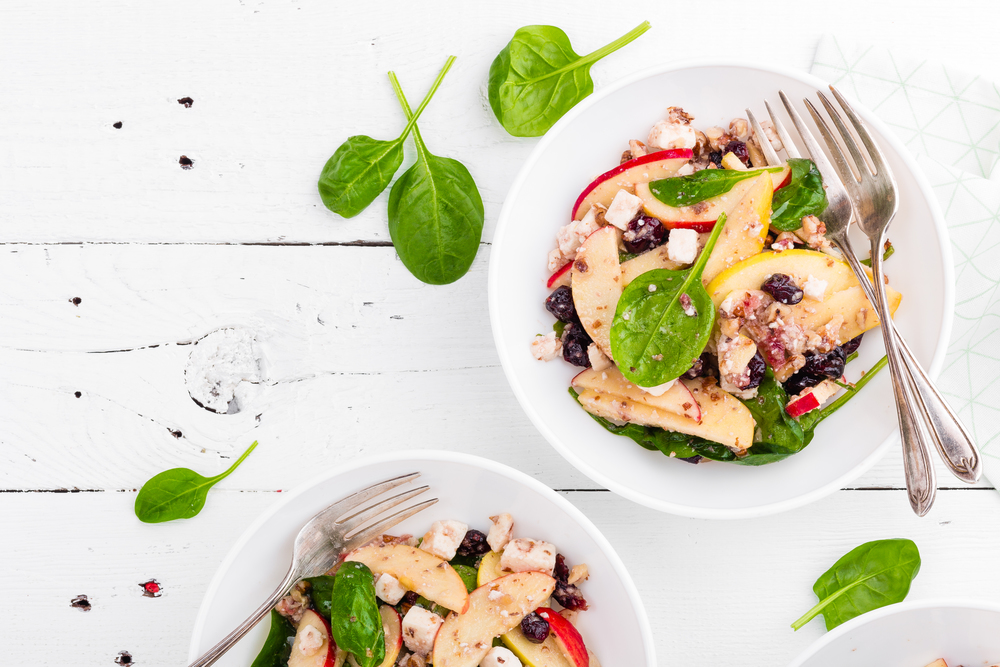 Healthy fruit and berry salad with fresh apples, cranberries, walnuts, italian ricotta cheese and spinach leaves. Delicious and nutritious diet dish for breakfast. Salad bowls on white wooden background. Overhead view