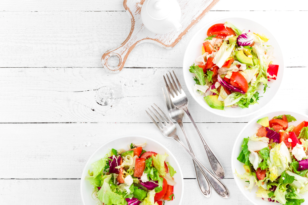 Healthy vegetable salad with fresh greens, lettuce, avocado, tomato, seet pepper and goat cheese. Delicious and nutritious diet dish for breakfast. Salad bowls on white wooden background. Top view