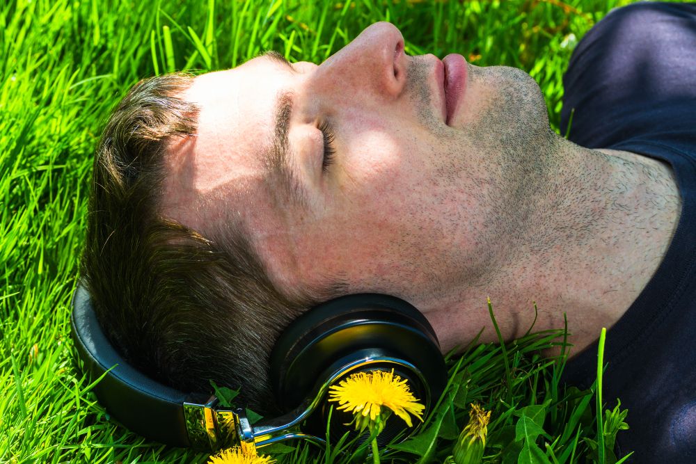 young man listening music with wireless headphones, lying on the lawn with dandelions