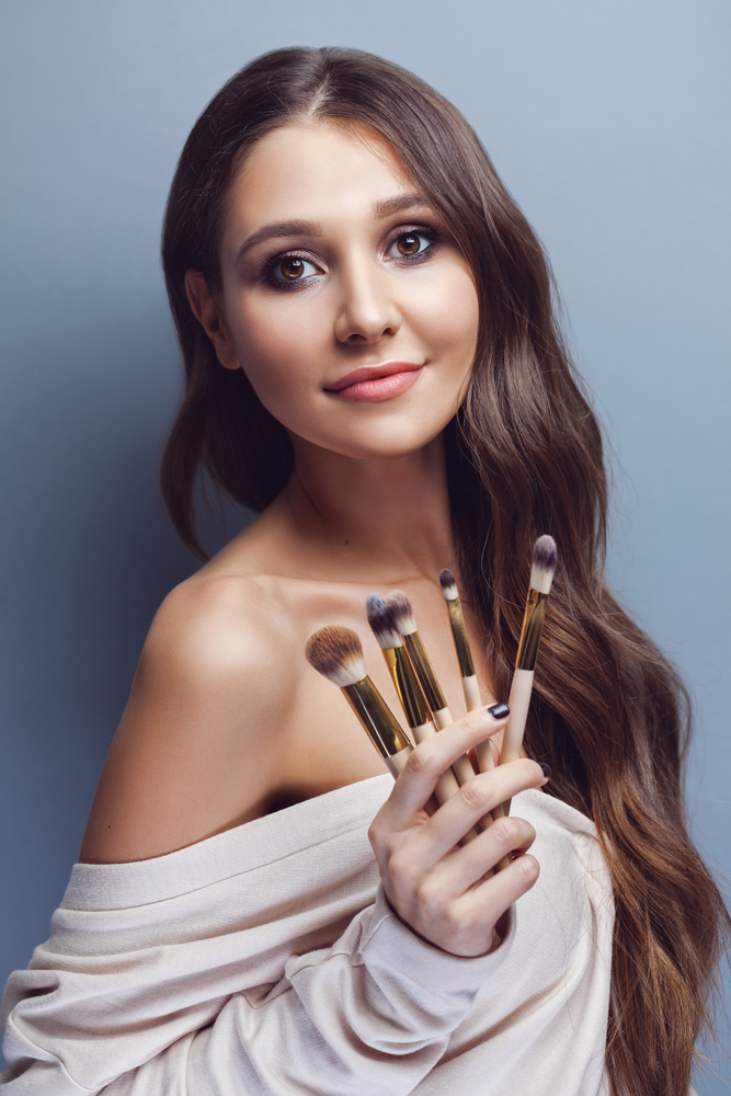 Beautiful young woman with long hair holding different make-up brushes, pastel blue background