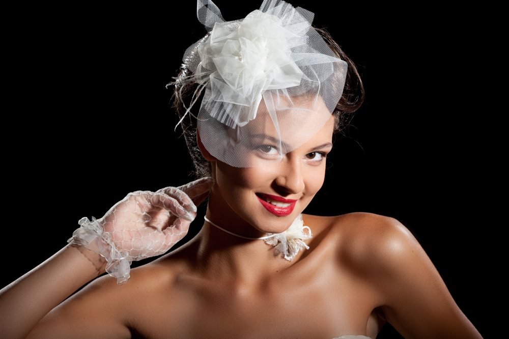 Portrait of Beautiful Elegant Bride in Wonderful Wedding Accessories: Lace Gloves,  White Necklace and Fancy Hat at the Black Background.