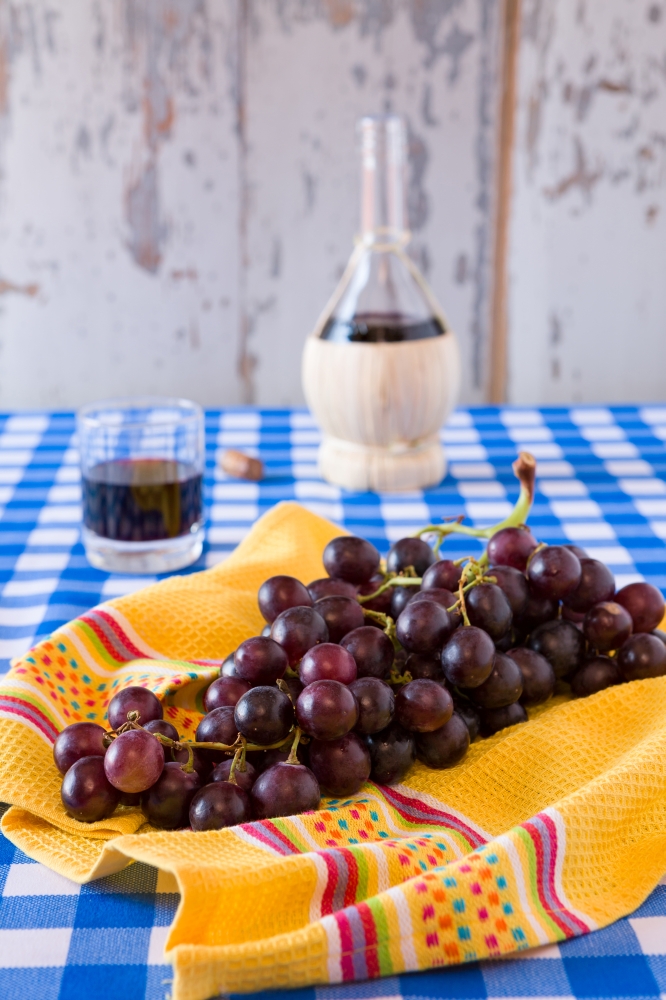 Bunch of red grapes and a glass of red wine with a wine flask on background over a checkered tablecloth. Bunch of red grapes and a glass of red wine with a wine flask
