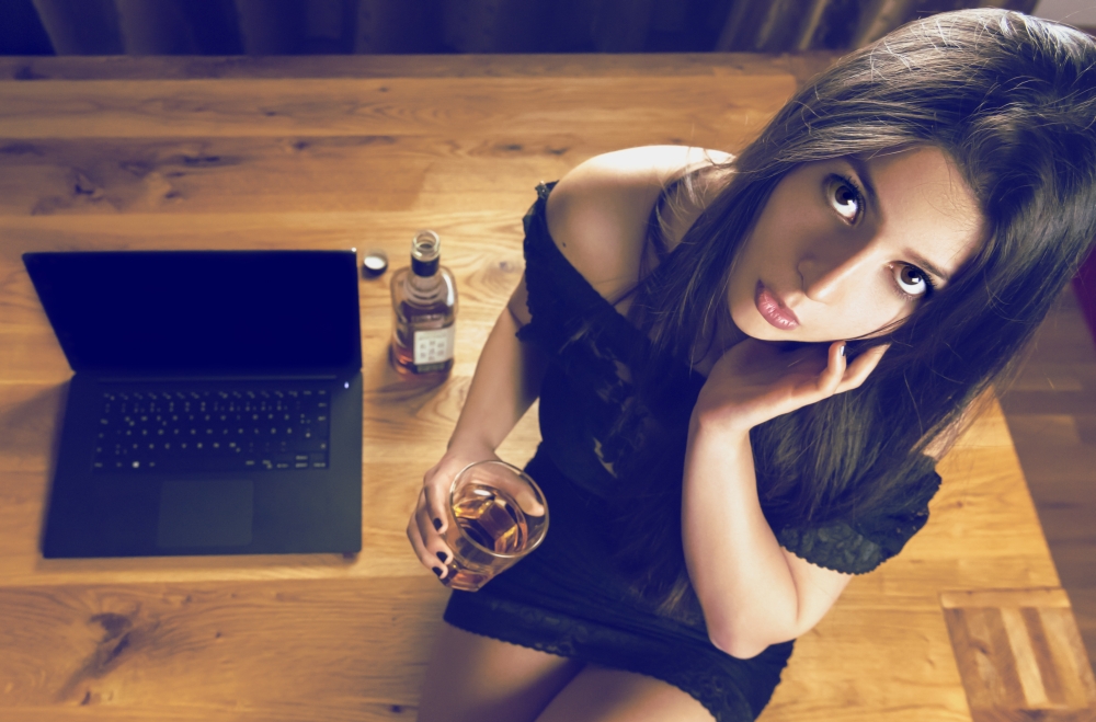 Young woman wearing a sexy black dress, holding a glass of whiskey in hand and sitting near the laptop on a wooden office