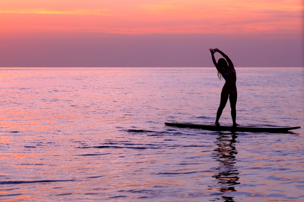 Silhouette of a girl practicing yoga, balancing on the surf board in the sea over beautiful purple sunset, healthy lifestyle and active summer vacation