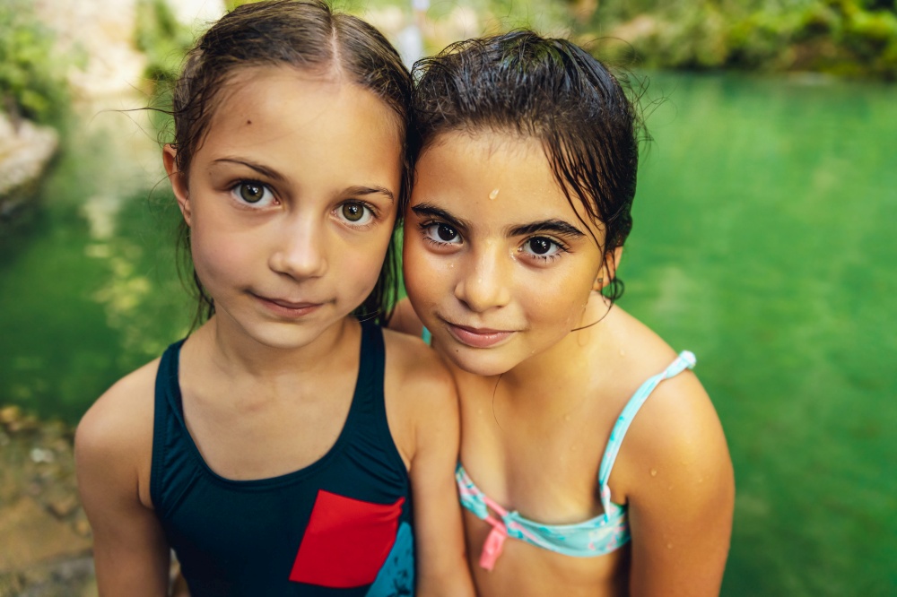 Portrait of a two cute little girls standing near the mountain&rsquo;s lake, with pleasure spending time in summer camp, happy best friends enjoying summer time together