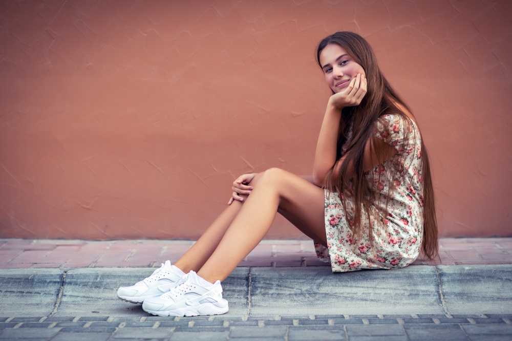 Pretty girl on the street, nice female with long natural hair sitting on blank monophonic wall background, authentic genuine beauty of young people