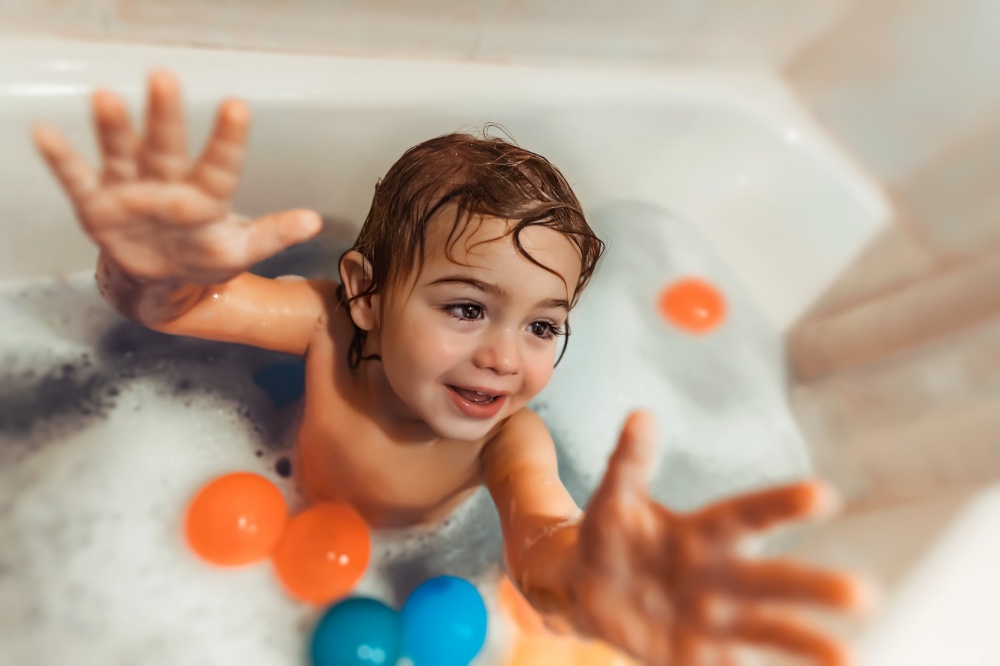 Cheerful boy taking bath, little child with pleasure bathing with foam and toys, happy healthy childhood concept