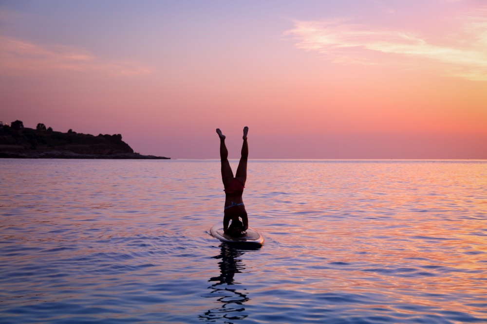 Silhouette of a sportive woman doing yoga exercise on the sup board over sunset, doing Sirsasana, headstand, active lifestyle, enjoying water sport, active summer vacation