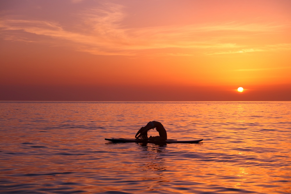 Silhouette of a yoga trainer balancing on the water on the paddle board over beautiful orange sunset background, doing yoga asana Urdhva dhanurasana on the beach