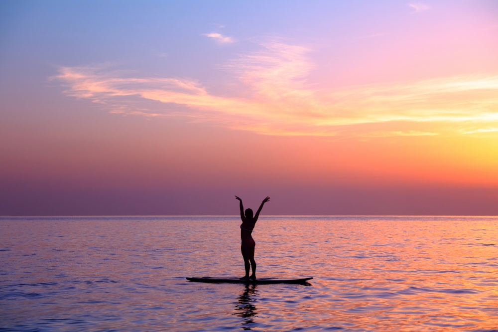 Silhouette of a yoga trainer over sunset background doing asanas, balancing on the sup board, enjoying healthy lifestyle, summer vacation on the beach