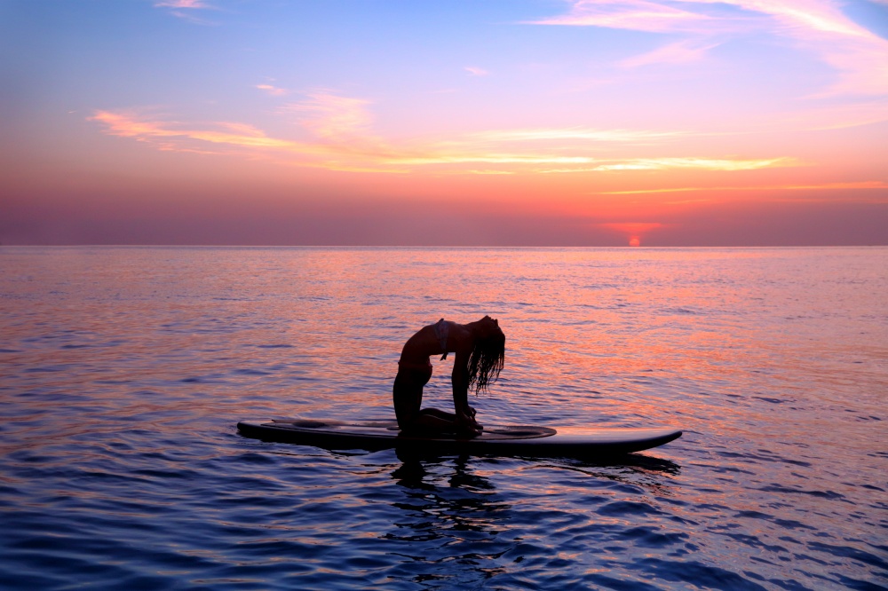Silhouette of a yoga trainer balancing on the water on the paddle board over beautiful purple sunset background, doing yoga asana Urdhva dhanurasana on the beach