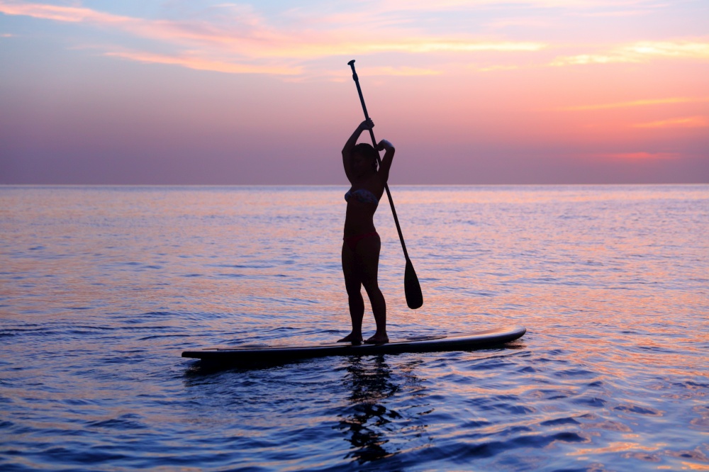 Silhouette of a girl practicing SUP, balancing on paddle board in the sea over beautiful purple sunset, healthy lifestyle and active summer vacation