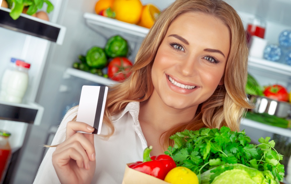 Buyer of a healthy food, portrait of a nice female with a credit card and a paper bag full of fresh vegetables, standing near open fridge at home, organic nutrition and weight loss concept