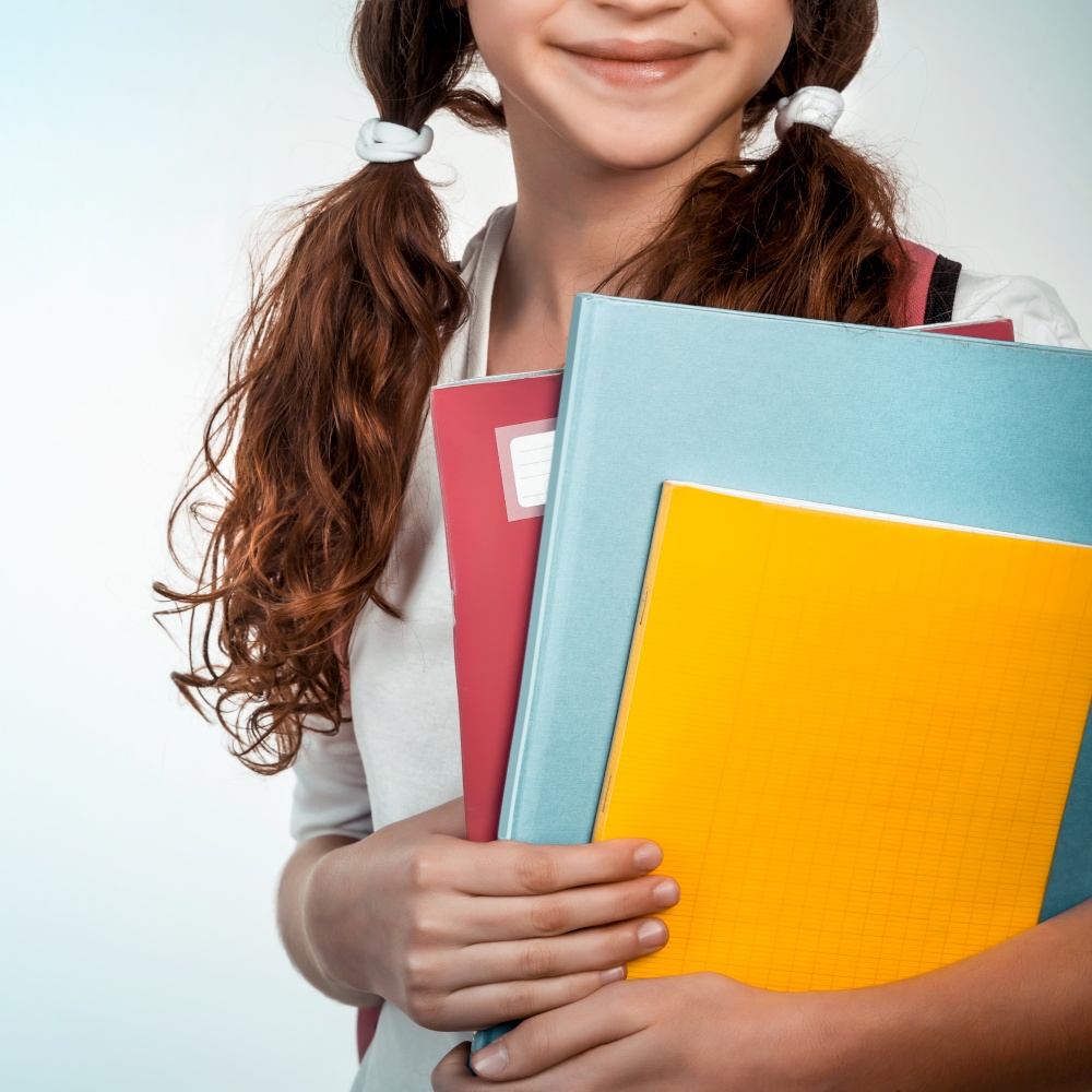 Portrait of cute brunette schoolgirl holding in hands three colorful notebooks, face part, isolated on clear background, back to school concept