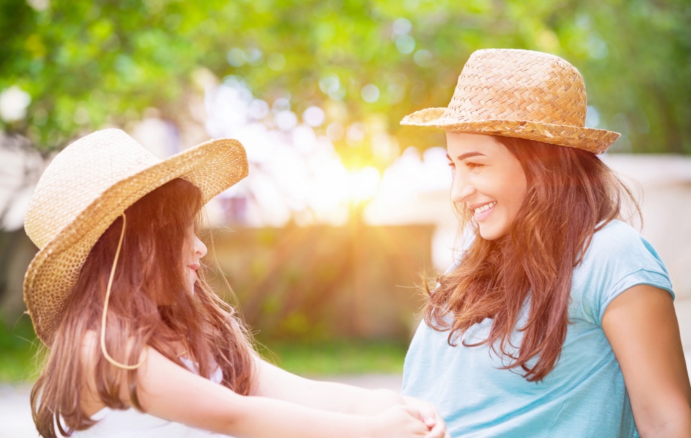 Portrait of a joyful young mother with her cute cheerful daughter wearing same straw hats and playing outdoors, laughing and looking on each other, portrait of a happy family enjoying life