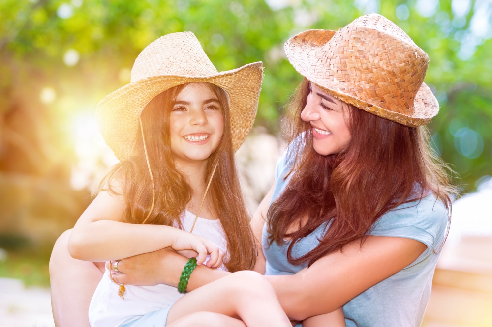Portrait of a joyful young mother with a cute cheerful daughter wearing same straw hats and playing outdoors, laughing and having fun, portrait of a happy family enjoying life