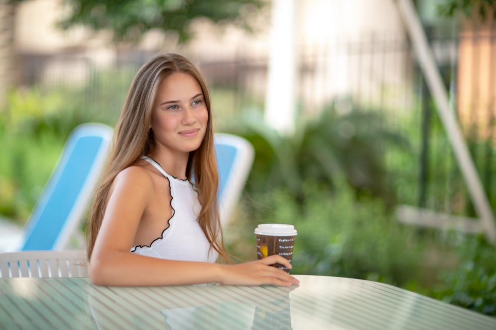 Cute female sitting outdoors with cup of coffee, having strong energy drink for breakfast, spending peaceful summer holidays on the beach resort