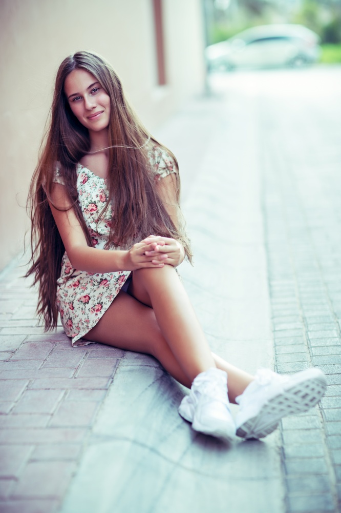 Nice teen girl wearing casual clothes and sitting on the sidewalk on the street, spending leisure time outdoors, genuine beauty of young people