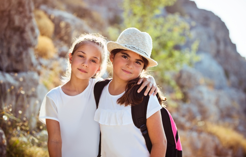 Portrait of a two cute school girls outdoors with backpacks, spending holidays in summer camp, happy active childhood