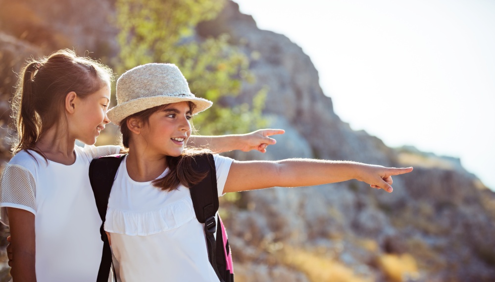 Two happy little girls enjoying excursion to the mountains, with pleasure discovering wild nature, with wonder looking on something and pointing, happy active time in a summer camp