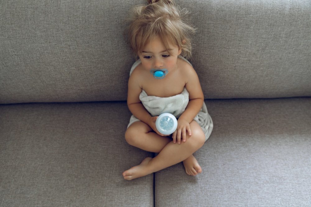 Sweet Adorable Child With Pacifier in the Mouth Sitting on the Sofa at Home Holding Baby Bottle with Milk. Time to Eat. Happy Healthy Childhood.
