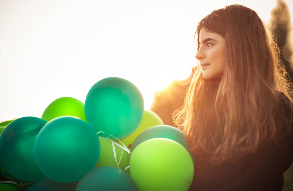 Portrait of a cute girl outdoors with big pile of green air balloons, preparing to celebrate birthday party, happy carefree childhood