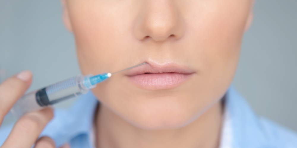 Closeup Photo of a Women&rsquo;s Face Part with Syringe over Gray Background. Lip Augmentation Procedure. Nice Female in a Beauty Clinic.. Woman Making a Lip Augmentation