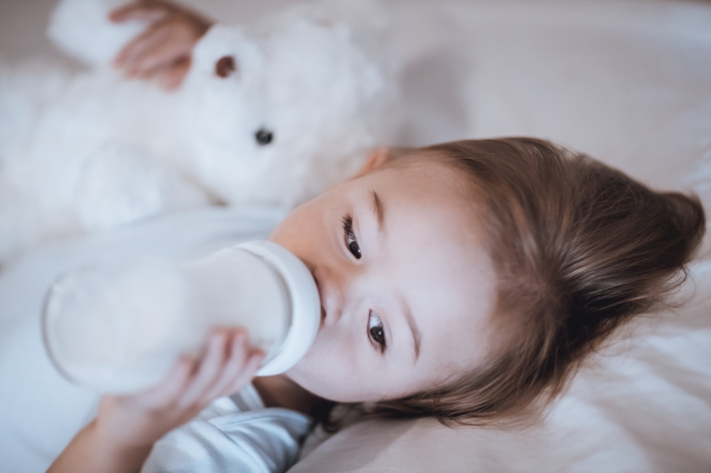 Portrait of a Cute Little Baby with Pleasure Drinking Formula Before Nap. Hugging Nice White Teddy Bear. Happy Healthy Childhood.. Sweet Baby Drinking Formula